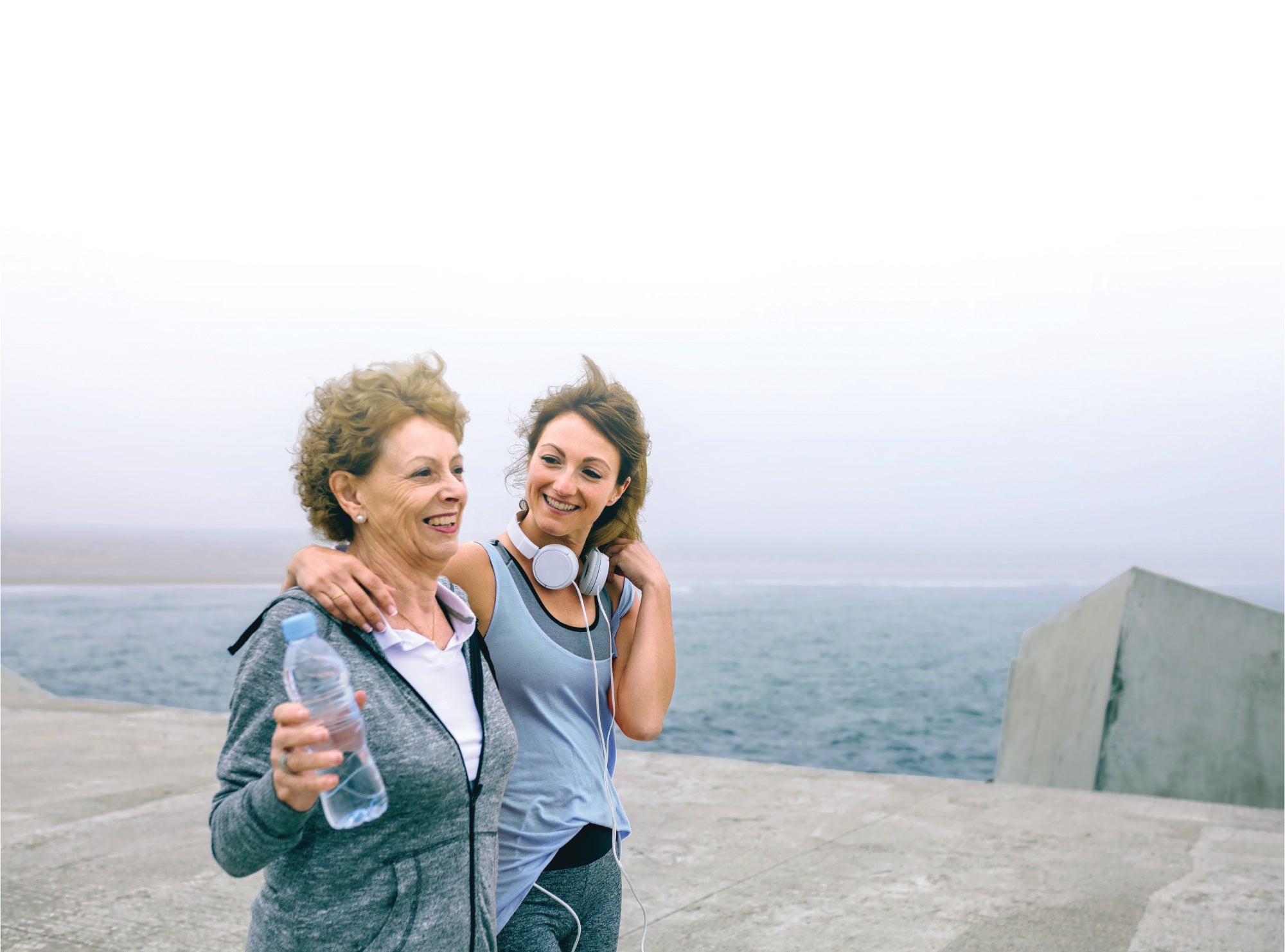 Two women smiling, one holding a water bottle, both in workout attire, walk together by the sea on a cloudy day.