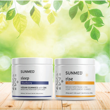 Two jars of Sunmed CBD Broad Spectrum Sleep + Rise Gummies Bundle are placed on a wooden surface with a leafy, sunlit background.