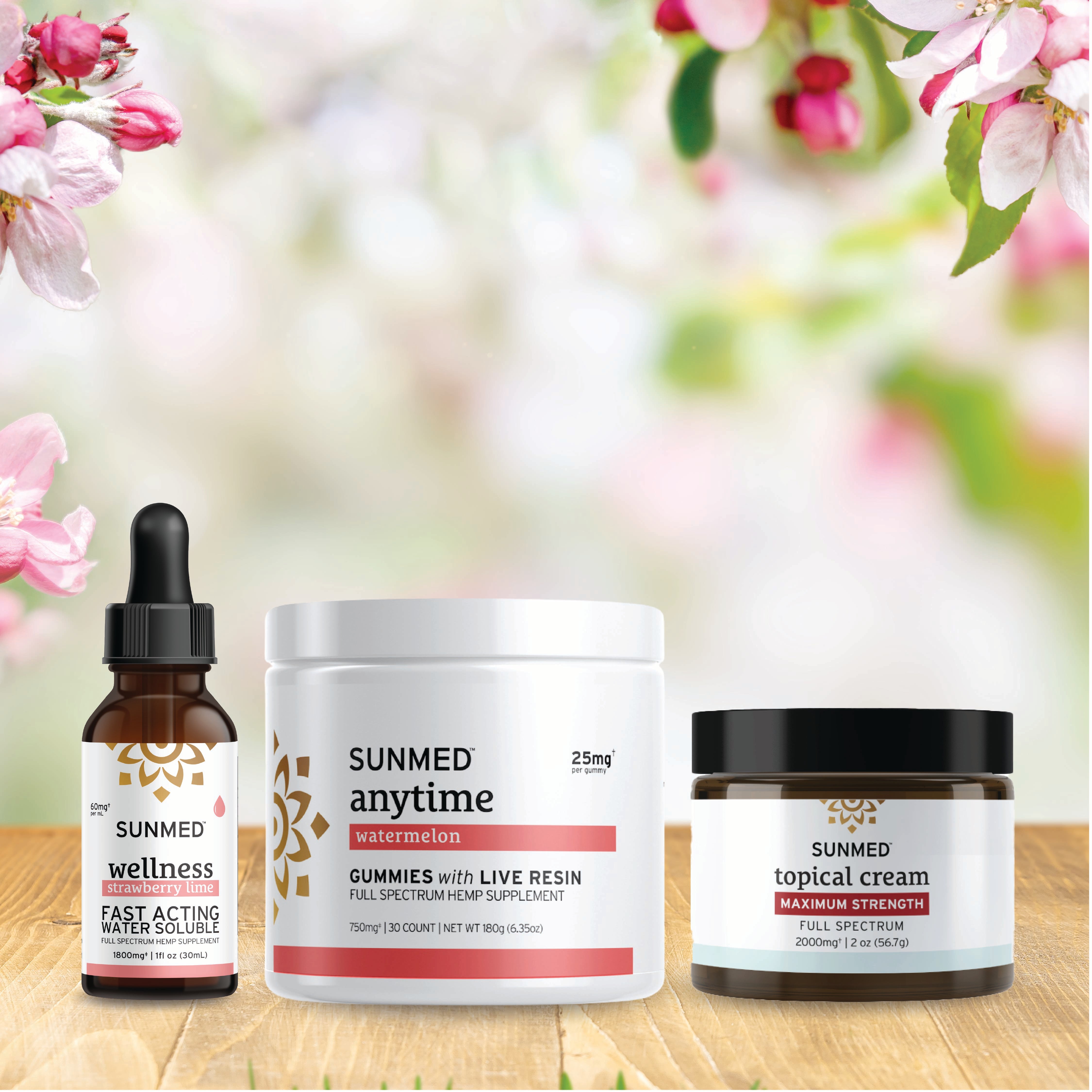 Three Sunmed CBD products—a Full Spectrum Anytime Gummies + Water Soluble + Topical Cream Bundle—are displayed on a wooden surface, accented by pink and white flowers in the background.