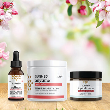 Three Sunmed CBD products—a Full Spectrum Anytime Gummies + Water Soluble + Topical Cream Bundle—are displayed on a wooden surface, accented by pink and white flowers in the background.