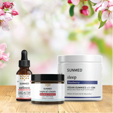 Three Sunmed CBD wellness products from the Broad Spectrum Sleep Gummies + Water Soluble + Topical Cream Bundle are displayed on a wooden surface, with pink and white flowers in the background.