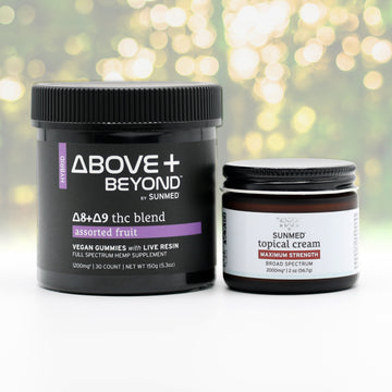 Two black jars from Sunmed CBD’s ABOVE + BEYOND Gummies + Topical Cream Bundle rest on a white surface, offering a soothing duo with nature-inspired relief, set against blurred greenery.