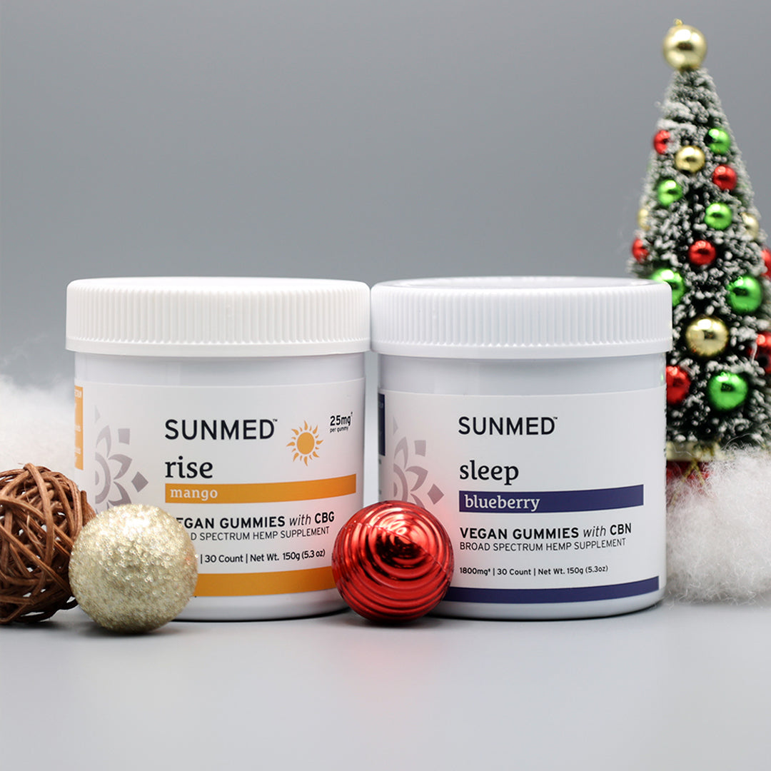 Two jars of Sunmed CBD's Broad Spectrum Sleep + Rise Gummies Bundle with holiday decor sit by a small Christmas tree, bringing THC-free wellness to your festive season.