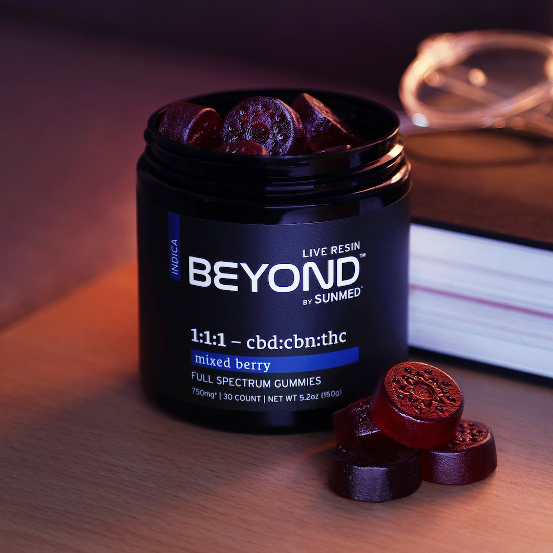 A jar of Sunmed CBD's Beyond 1:1:1 Indica Live Resin Gummies, featuring a premium mixed berry flavor, sits invitingly next to a book and eyeglasses in the background.