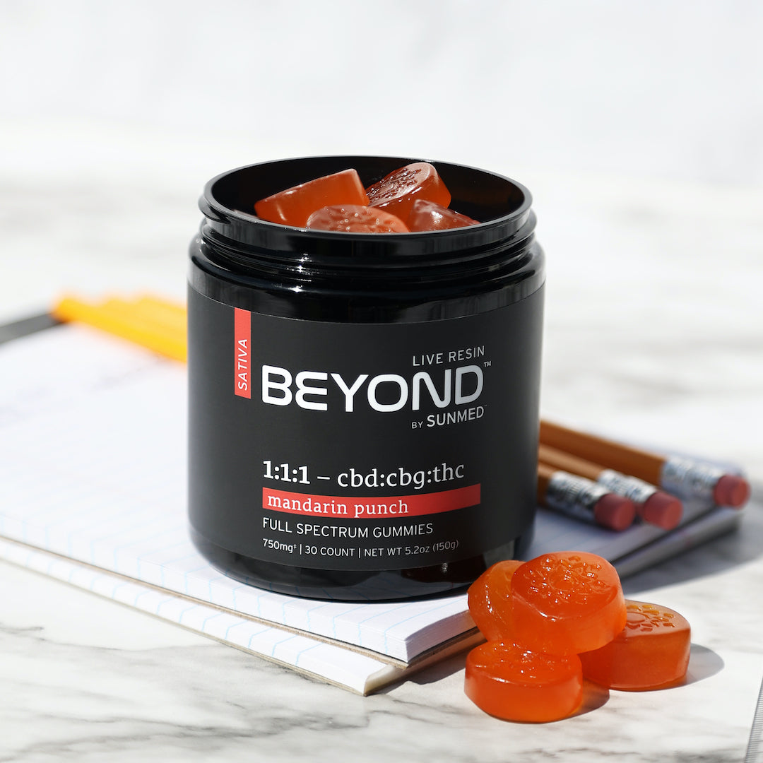 A jar of Sunmed CBD’s Beyond 1:1:1 Sativa Live Resin Gummies featuring a mandarin punch flavor, placed next to some gummies and pencils on a notepad, offering a creative energy boost.