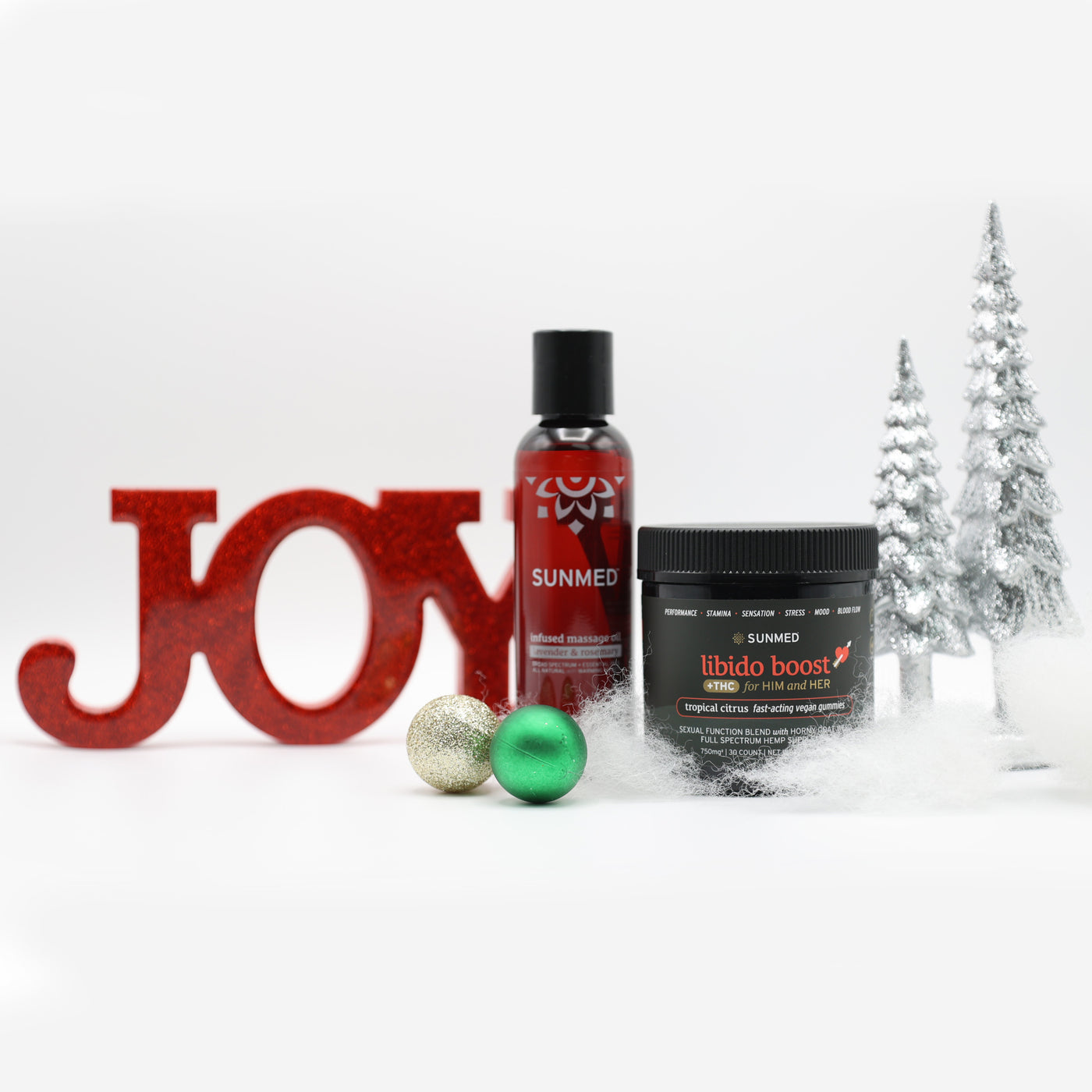 A red "JOY" sign, Sunmed CBD's Libido Gummies THC + Massage Oil Bundle, green ornaments, and silver trees are arranged on a snowy white background.