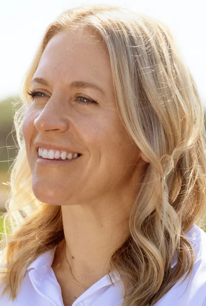 Smiling woman in a white SUNMED polo stands outdoors with blurred greenery in the background.