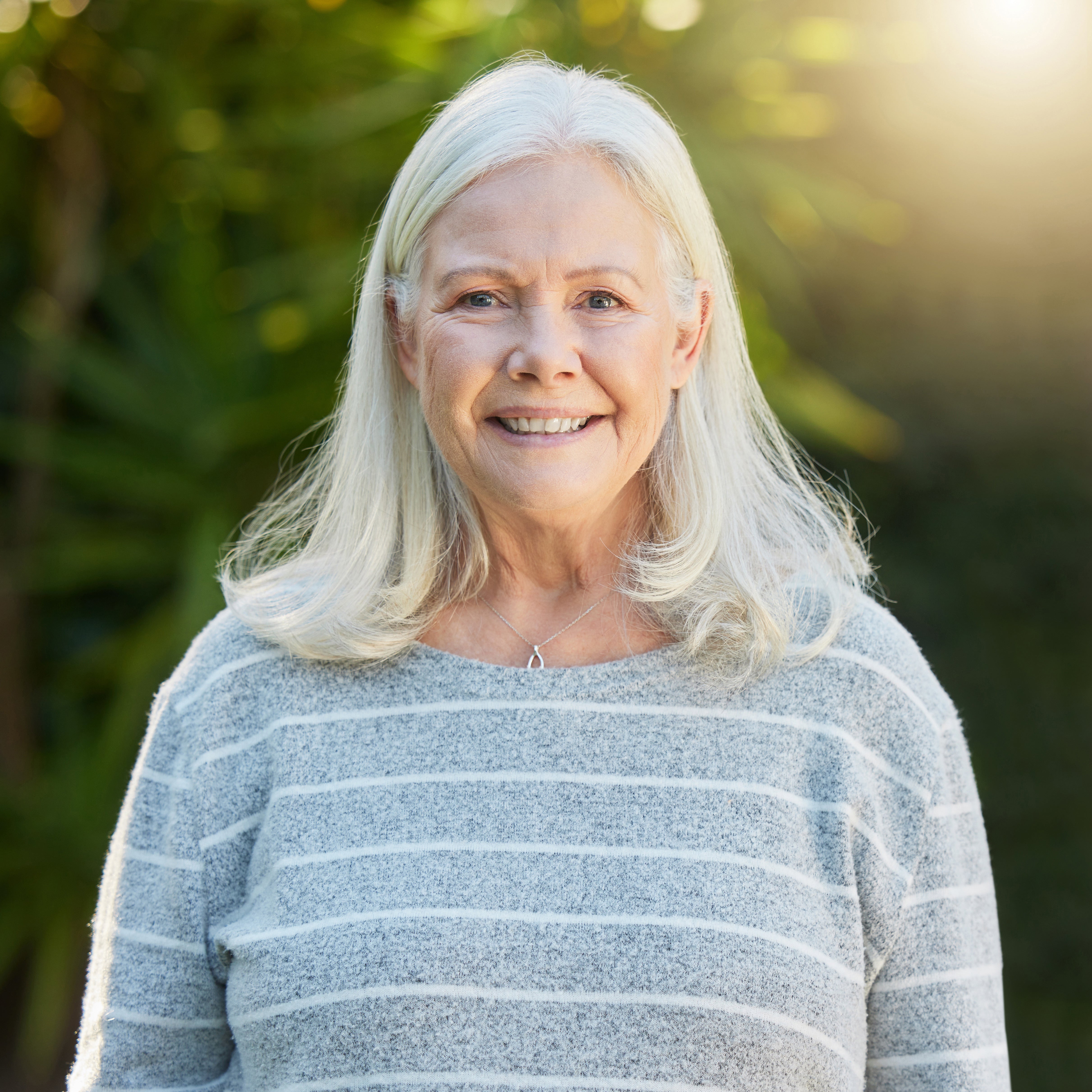 Smiling older woman with gray hair and striped sweater stands outdoors in sunlight, looking well-rested after using Sunmed CBD’s Full Spectrum Sleep CBN Gummies to improve her sleep quality.