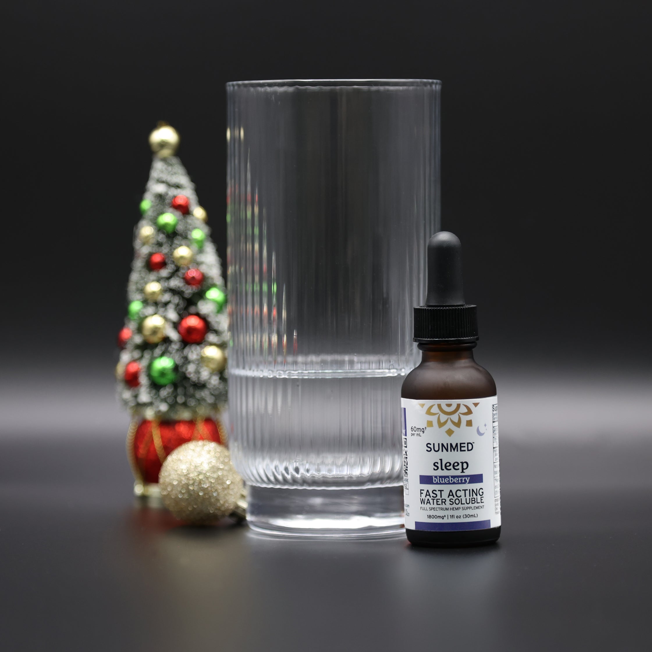 A glass of water, a bottle of Sunmed CBD Full Spectrum Sleep CBN Water Soluble with fast-acting hemp extract, and a small decorated Christmas tree on a black background.