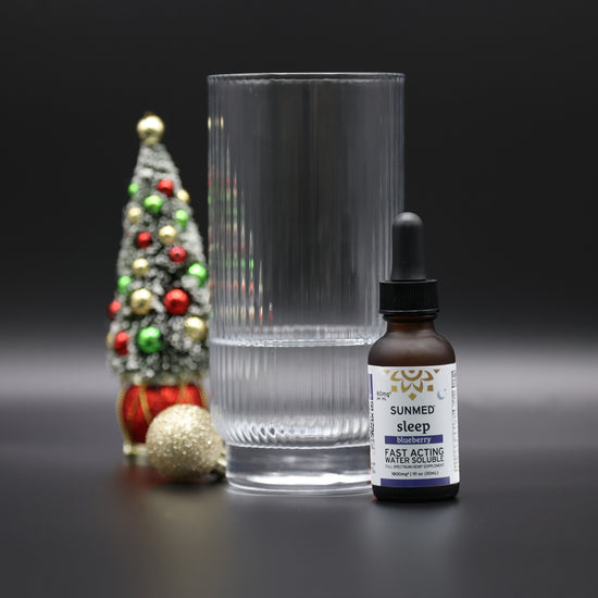 A glass of water, a bottle of Sunmed CBD Full Spectrum Sleep CBN Water Soluble with fast-acting hemp extract, and a small decorated Christmas tree on a black background.