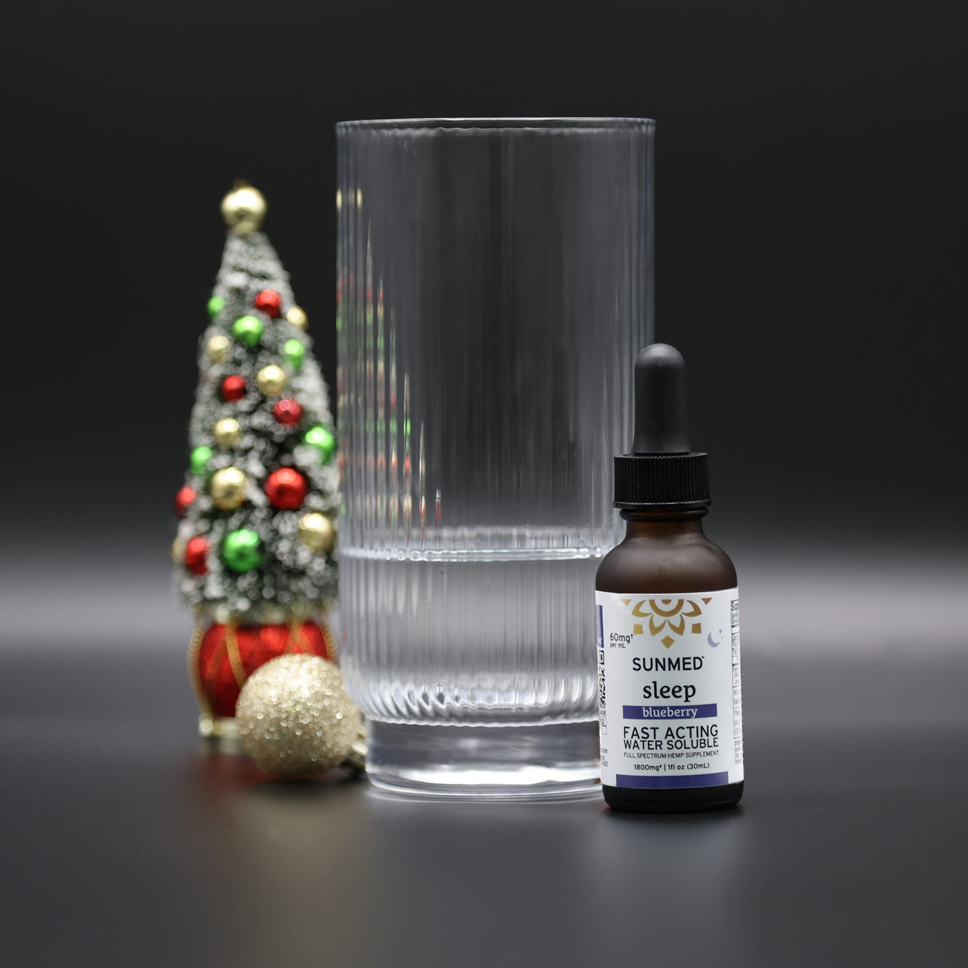 A glass of water, a bottle of Sunmed CBD Full Spectrum Sleep CBN Water Soluble with fast-acting hemp extract, and a small decorated Christmas tree on a black background.