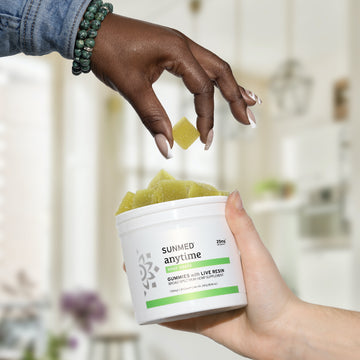 Close-up of a hand picking a Full Spectrum Anytime CBD Gummy from a Sunmed CBD container, filled with premium hemp extract, held by another hand in a kitchen setting.