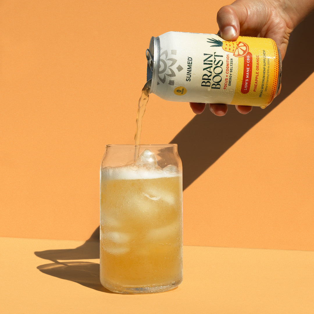 A hand pours Sunmed CBD's Brain Boost Energy Seltzer - 24 Pack from a can into a glass filled with ice against an orange background, infusing every sip with the power of lion's mane and broad spectrum hemp extract.