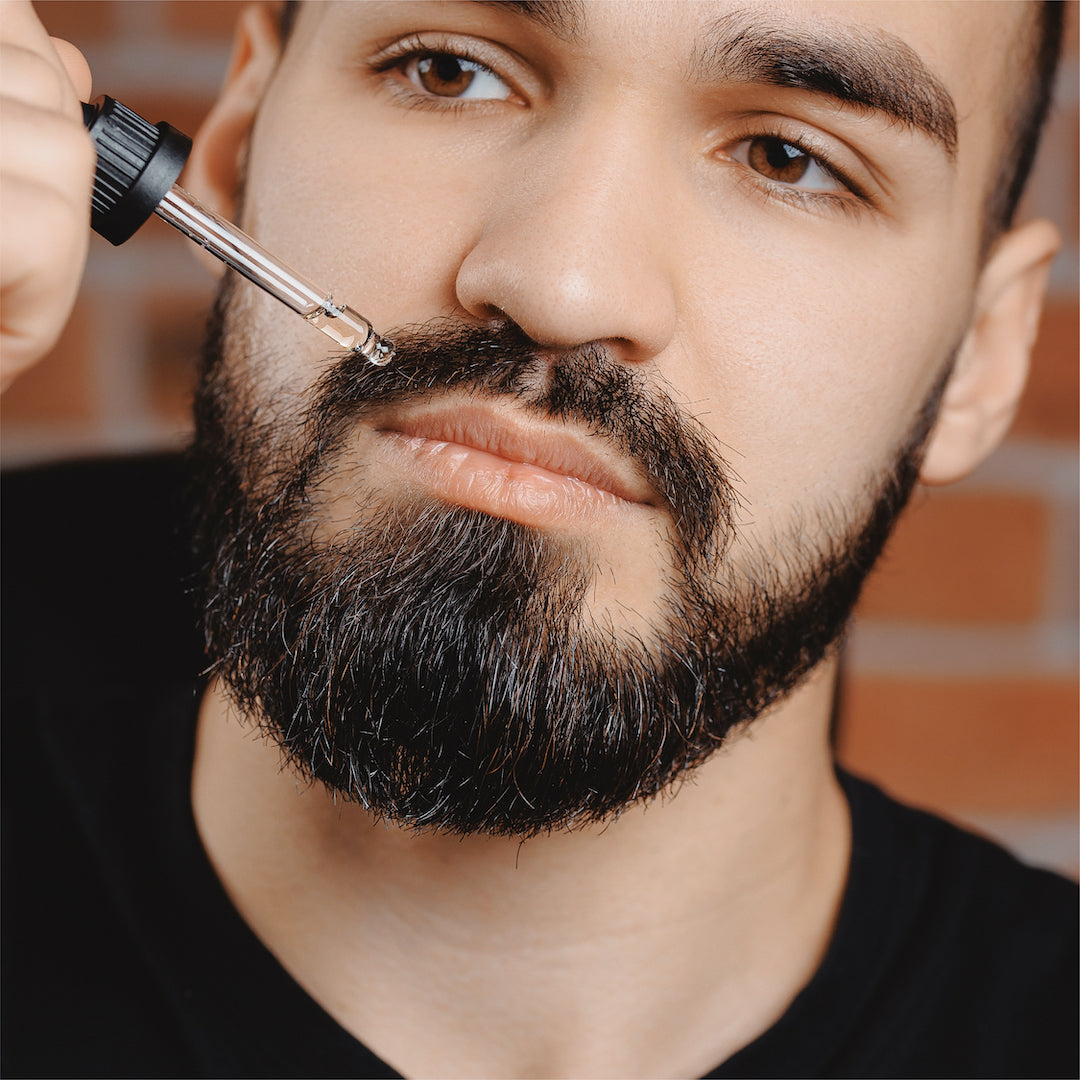 A man applying Sunmed CBD Broad Spectrum Hair + Scalp Oil with a dropper to his mustache, with a brick wall in the background.