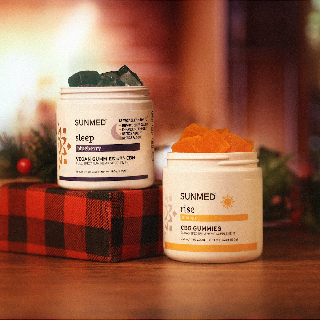 Two jars of the Sleep + Rise Gummies Holiday Bundle from Sunmed CBD are placed on a red plaid surface, surrounded by festive greenery.