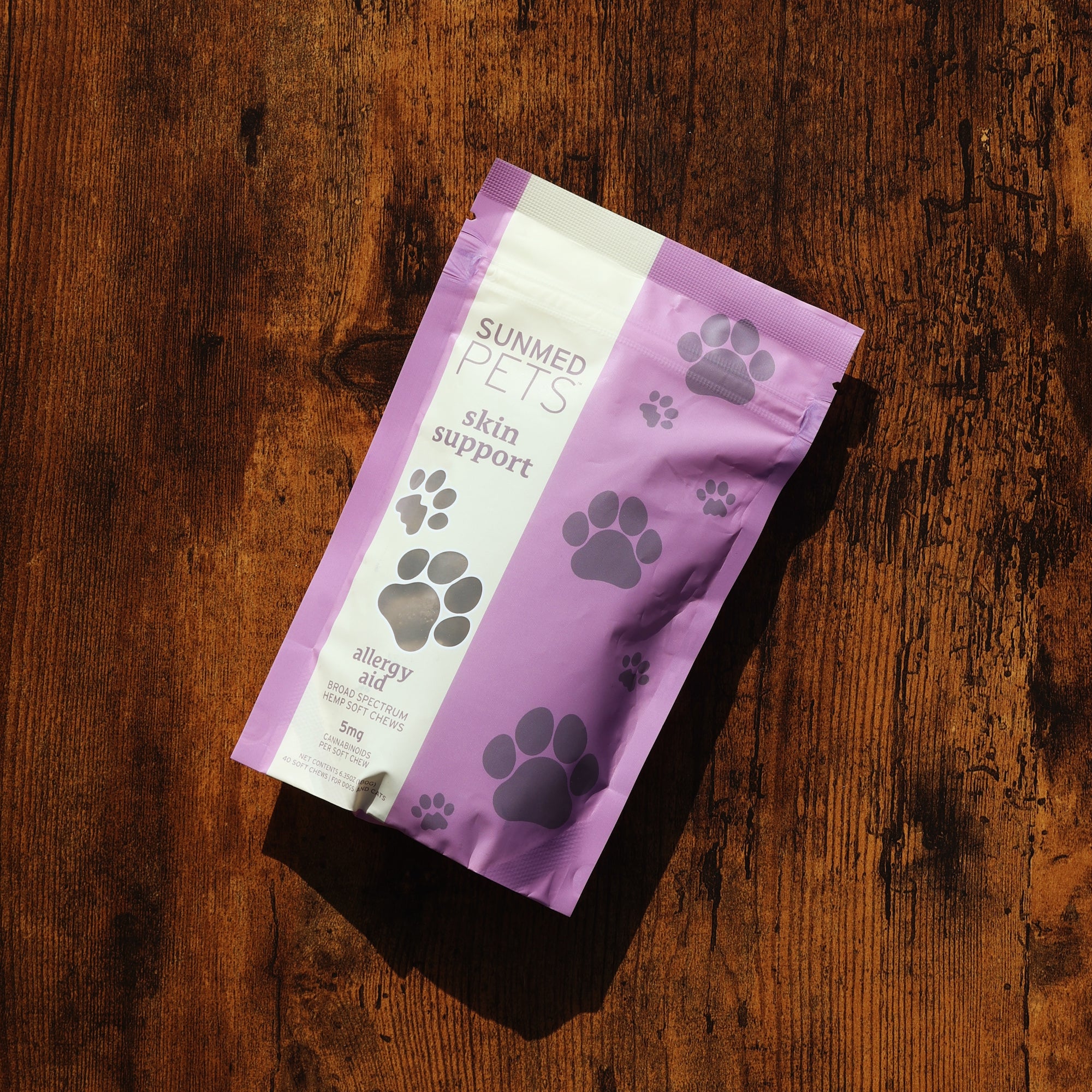 A purple Sunmed CBD Pets skin support bag with a paw print design, featuring Broad Spectrum Allergy Aid CBD Pet Chews for optimal skin health, lies on a wooden surface.