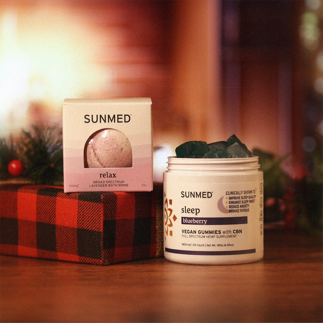 Sunmed CBD's Sleep Gummies + Bath Bomb Holiday Bundle displayed on a festive plaid surface, surrounded by pinecones and greenery in the background.