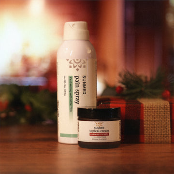 A couple of containers from the Menthol Spray + Topical Cream Holiday Bundle by Sunmed CBD rest on a wooden surface, accented with a festive touch in the background.