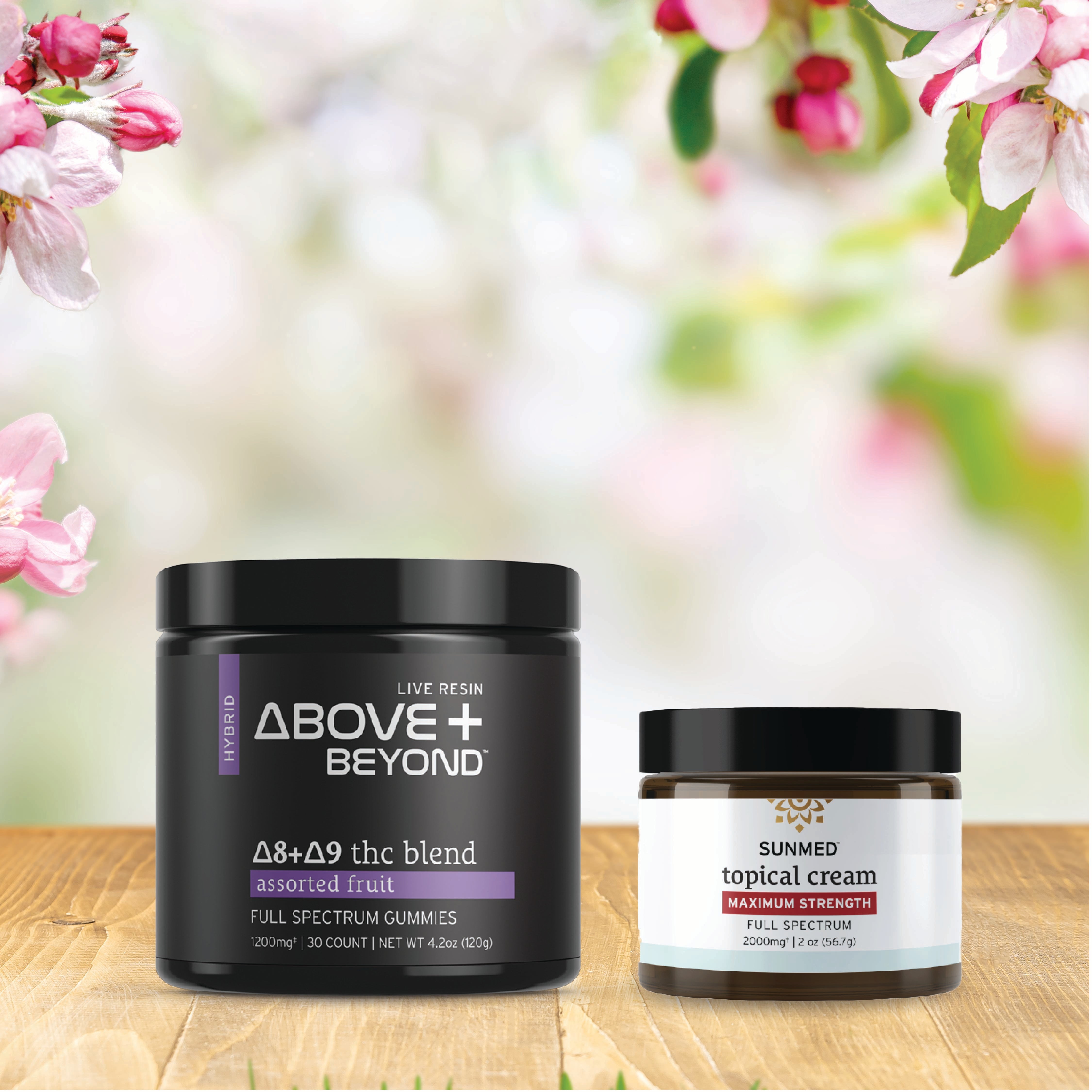 Two jars of Sunmed CBD’s ABOVE + BEYOND Gummies + Topical Cream Bundle sit on a wooden surface, with soft pink and white flowers blurred in the background.