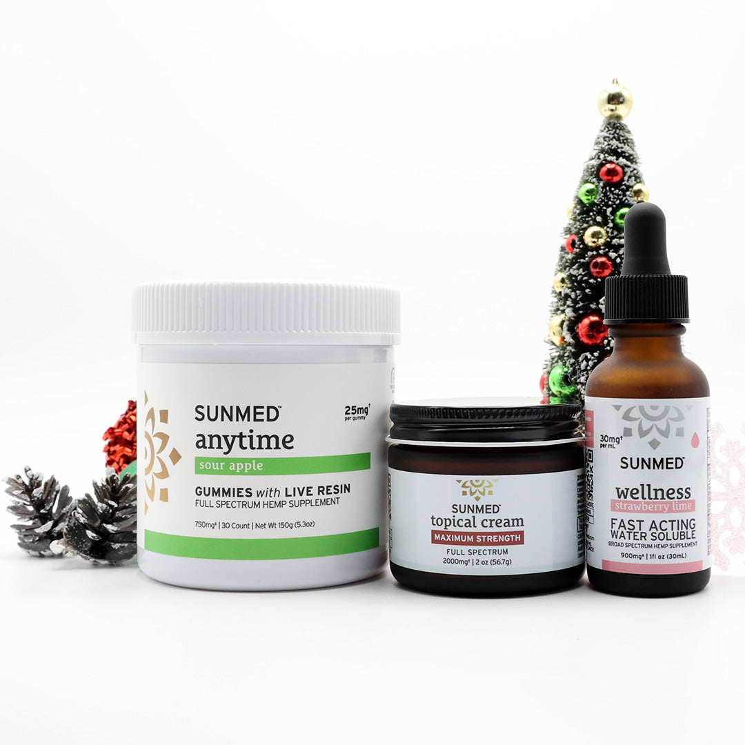 Three Sunmed CBD products—a Full Spectrum Anytime Gummies + Water Soluble + Topical Cream Bundle—are displayed in front of a small, decorated Christmas tree and pinecones on a white background.