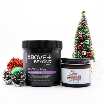 Two Sunmed CBD ABOVE + BEYOND Gummies + Topical Cream Bundle products are displayed with a decorated Christmas tree and festive ornaments in the background.