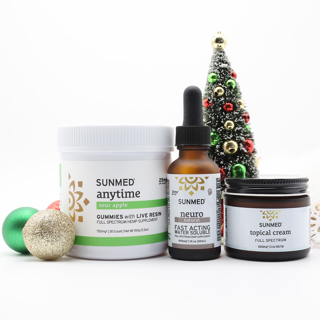 Three Sunmed CBD products—a Full Spectrum Anytime Gummies + Neuro Water Soluble + Topical Cream Bundle—are displayed in front of a decorated Christmas tree with ornaments on a white background.
