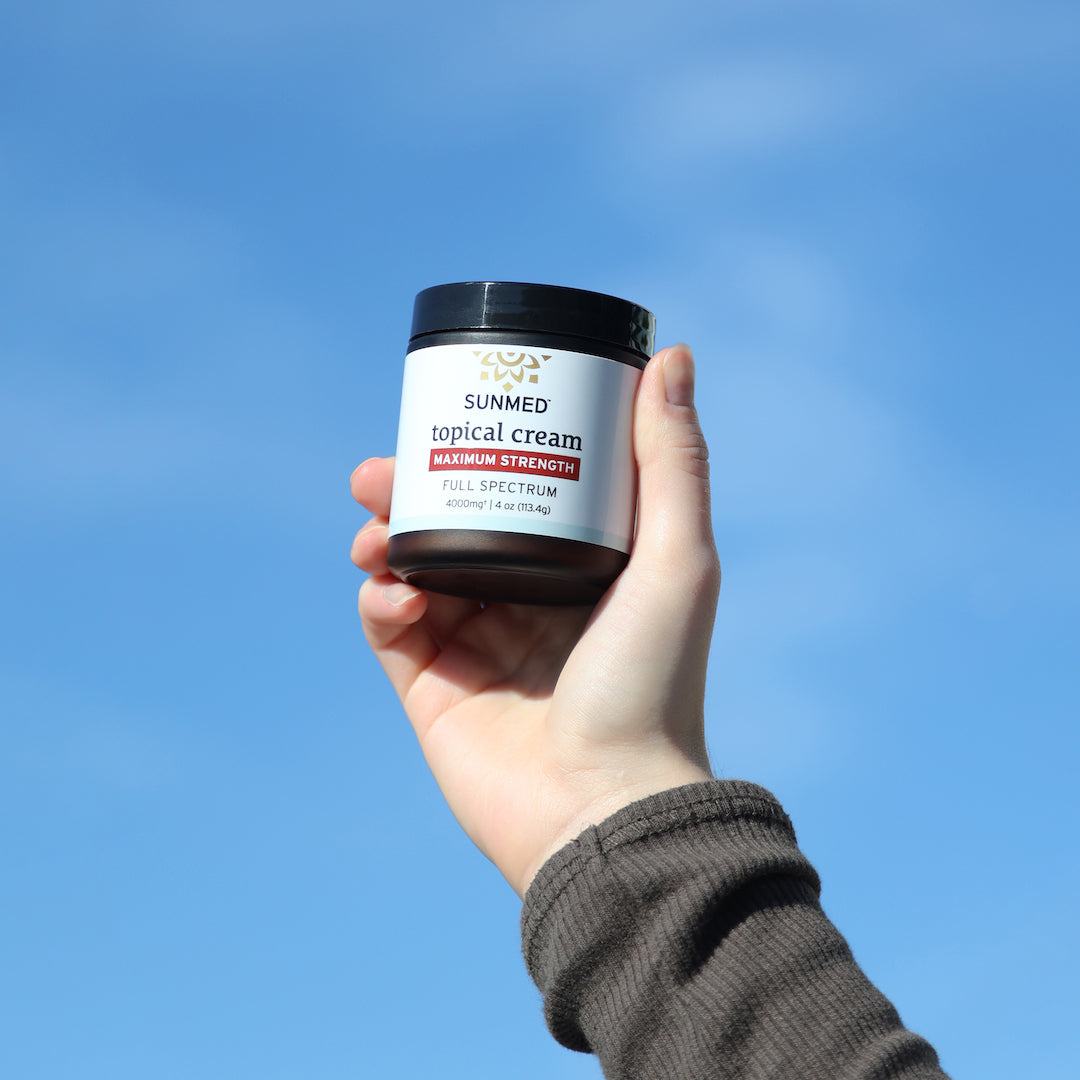 A hand holds a jar of Sunmed CBD Full Spectrum Relief CBD Cream against a clear blue sky background.