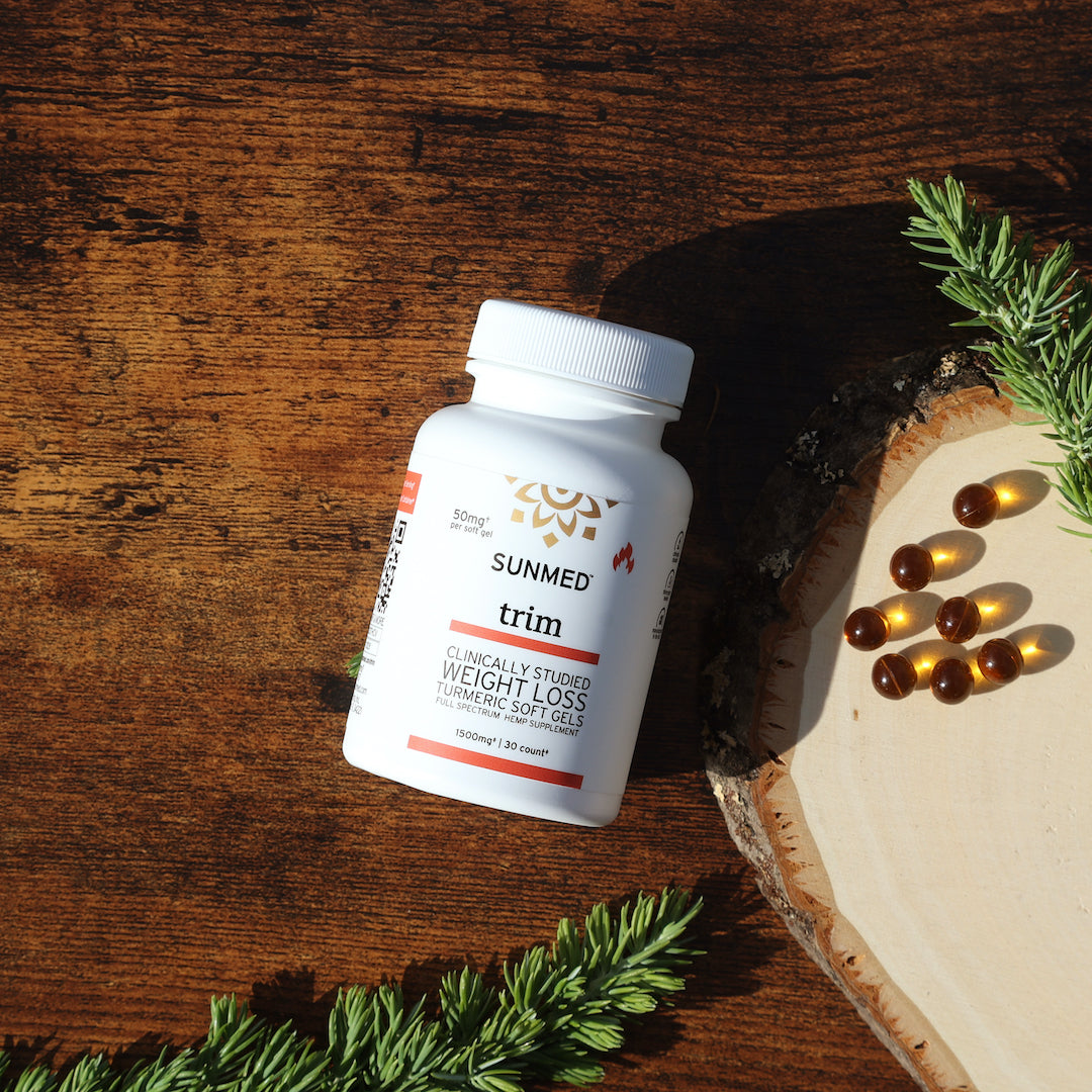 A bottle of Sunmed CBD Full Spectrum Trim Weight Loss Capsules, enriched with hemp extract and turmeric, sits on a wooden surface surrounded by several capsules and pine branches.