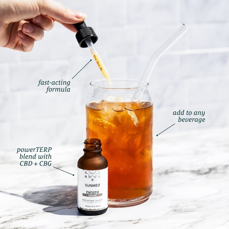 Hand holding dropper above iced tea glass, with labeled "Sunmed CBD Broad Spectrum Neuro CBD Water Soluble" product bottle reading "Fast-Acting Calm Focus" on marble surface.